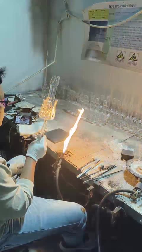 A worker manually handles a shaped glass component near a burner.